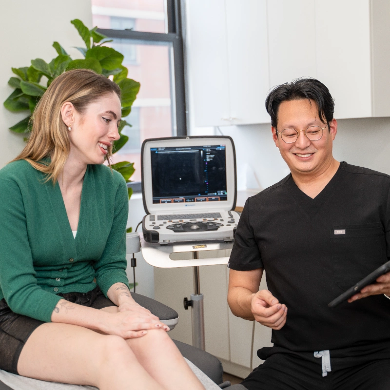 A friendly doctor in a white lab coat shows information about a vein treatment process on a tablet to a seated, smiling patient in a modern medical office setting.