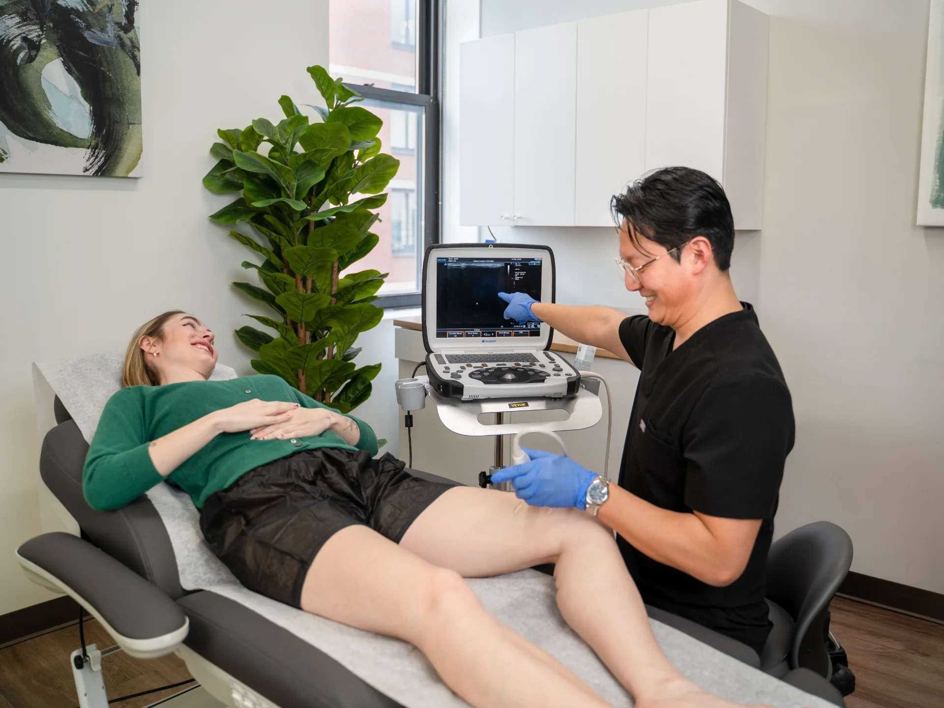 Dr. Benjamin Kum performs a duplex ultrasound examination on a patient's leg in a clinical setting. He uses an ultrasound probe on the patient's lower extremity while pointing at the ultrasound machine monitor to explain the diagnostic findings.