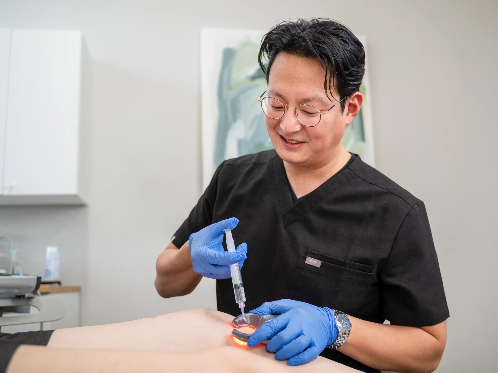 Dr. Benjamin Kum, wearing blue medical gloves and a wristwatch, uses a Veinlite to illuminate a patient's leg. He is carefully preparing to administer an injection with a syringe during the clinical procedure.