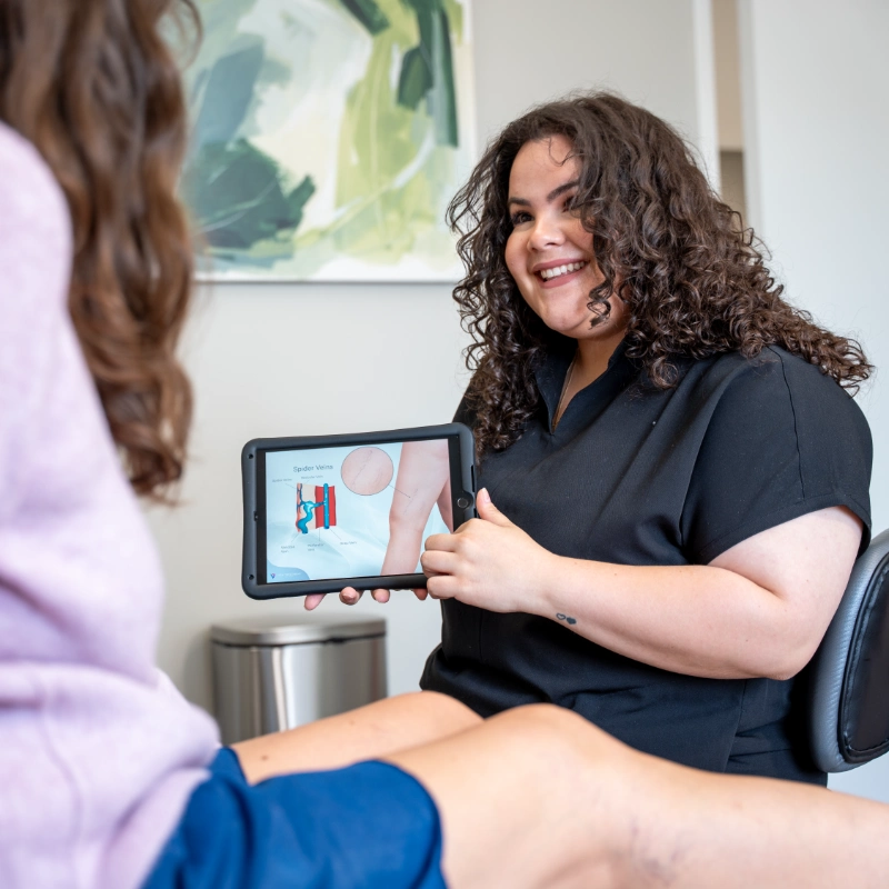 Dr. Hanen Fernandez consults with a patient in a clinical office setting, displaying educational content on a tablet device related to Spider Veins or Varicose Veins treatment options. The tablet shows diagnostic imaging or treatment planning information, facilitating patient education during the venous consultation in a professional medical environment with educational materials visible in the background.